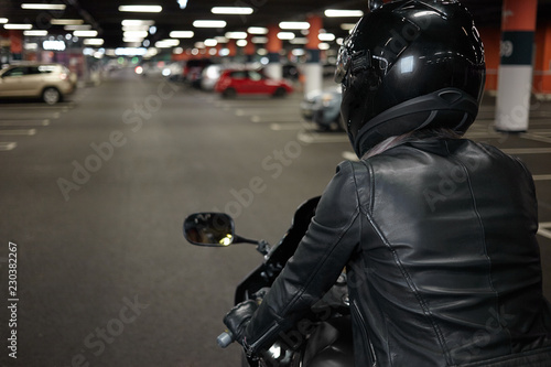 Rear view of unrecognizable woman wearing stylish motorcycle leather jacket, gloves and safety helmet, sitting in her two wheeled motor vehicle in underground garage or car parking, ready to drive