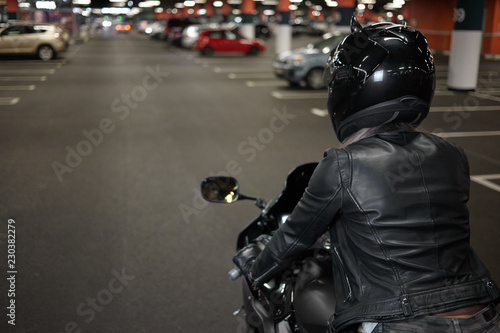 Active way of life, motorcycling, night city and people concept. Rear shot of fashionable confident female biker wearing safety helmet and black leather jacket, riding her motorbike on parking lot
