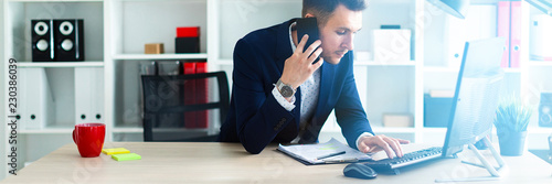 A young man is standing near a table in the office, talking on the phone and typing text on the keyboard. photo