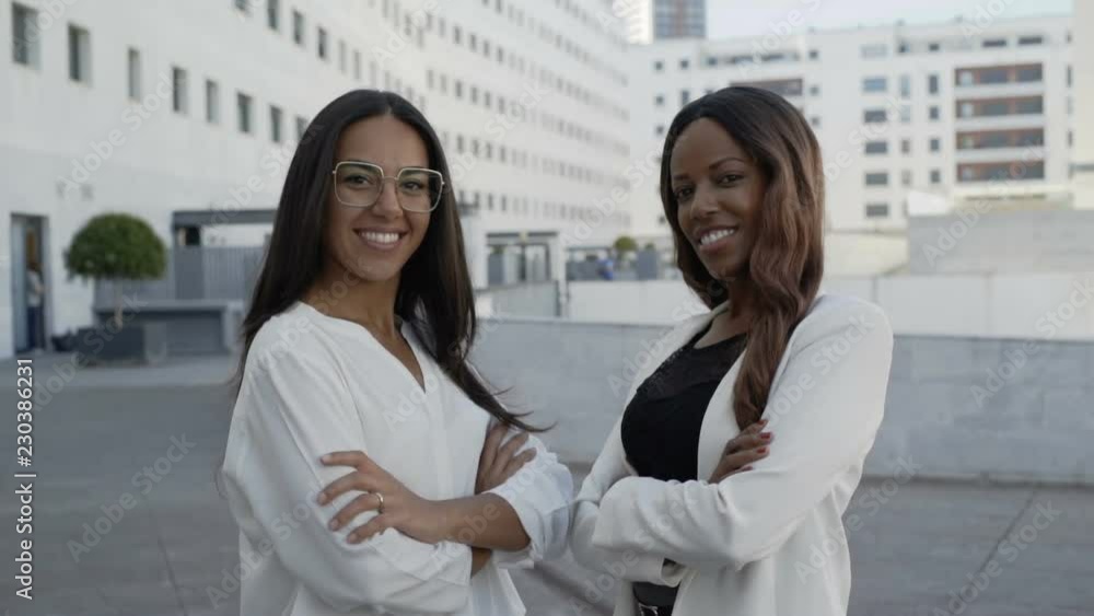 Multicultural successful partners posing with crossed arms near office building on street. Beautiful Latin and African American confident businesswomen standing together in urban city. Success concept Multicultural successful partners posing with crossed arms near office building on street. Beautiful Latin and African American confident businesswomen standing together in urban city. Success concept