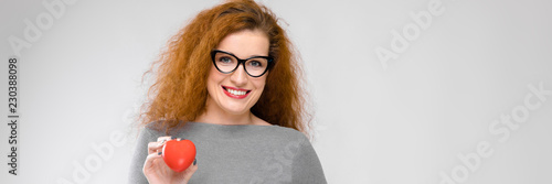 Charming young girl with red hair. A young girl in a gray sweater. Young girl on a gray background. A young girl with glasses is holding a red heart photo