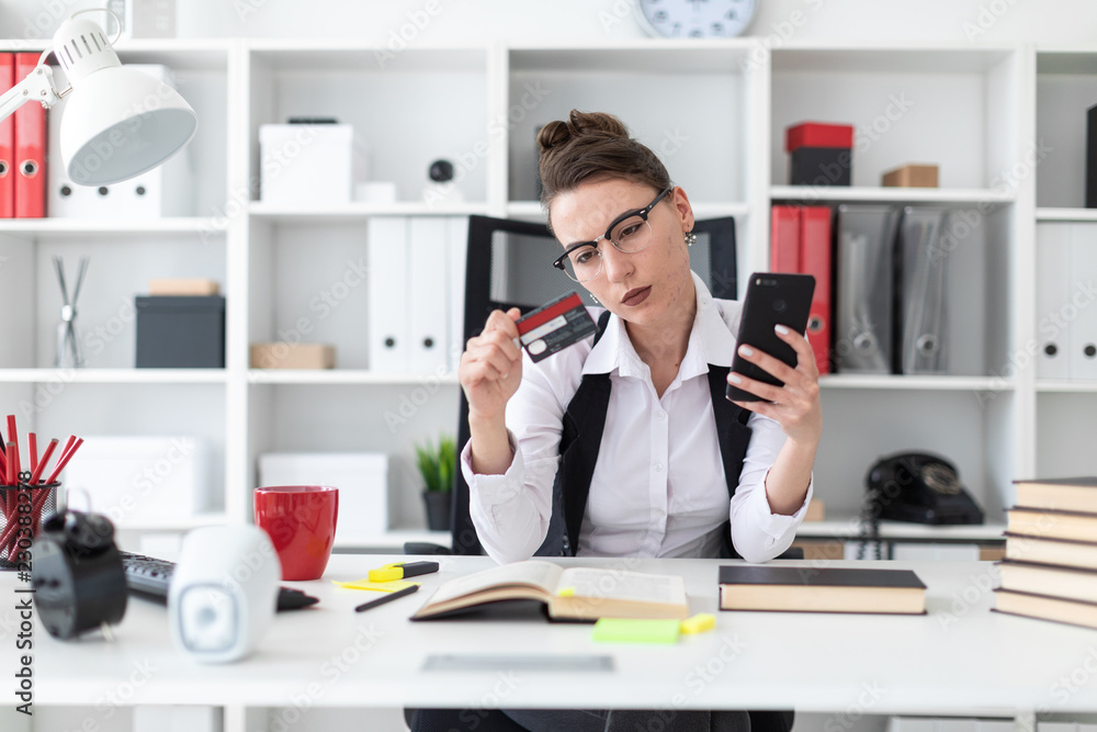 A young girl sits at a computer desk in the office and holds a bank card and phone. A young girl sits at a computer desk in the office and holds a bank card and phone.