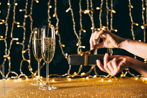 cropped image of woman opening christmas present near champagne in glasses on garland light background