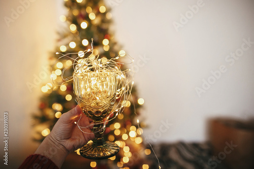 hand holding stylish vintage glass with garland bulbs inside on background of golden beautiful christmas tree with lights in festive room. cozy winter holidays. cheers for christmas.