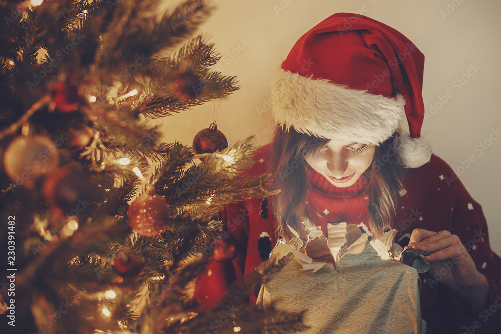 happy girl in santa hat opening magic Christmas gift box at golden beautiful christmas tree with lights and presents in festive room. winter atmospheric  moments. happy holidays.
