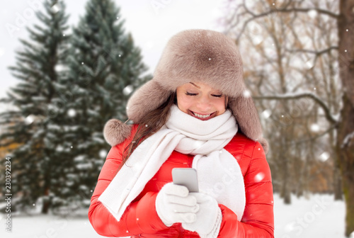 people, season and leisure concept - happy woman in fur hat with smartphone over winter forest background and show