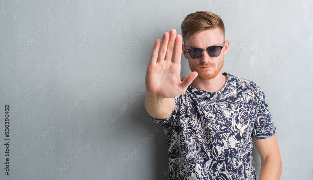 Young redhead man over grey grunge wall wearing sunglasses with open hand doing stop sign with serious and confident expression, defense gesture Young redhead man over grey grunge wall wearing sunglasses with open hand doing stop sign with serious and confident expression, defense gesture