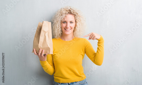 Young blonde woman over grunge grey background holding lunch paper bag with surprise face pointing finger to himself