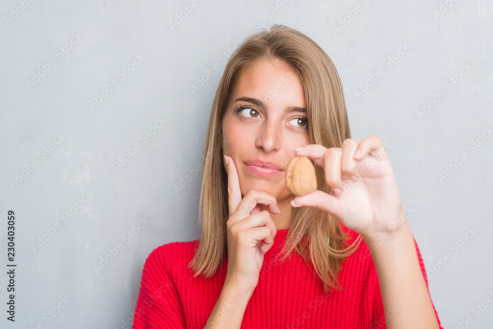 Beautiful young woman standing over grunge grey wall holding walnut serious face thinking about question, very confused idea Beautiful young woman standing over grunge grey wall holding walnut serious face thinking about question, very confused idea