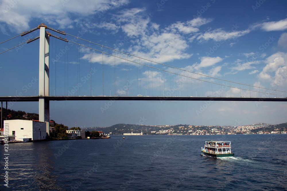 View of a tour boat with unrecognizable people on it, Bosphorus bridge. It is a sunny summer day with light clouds.