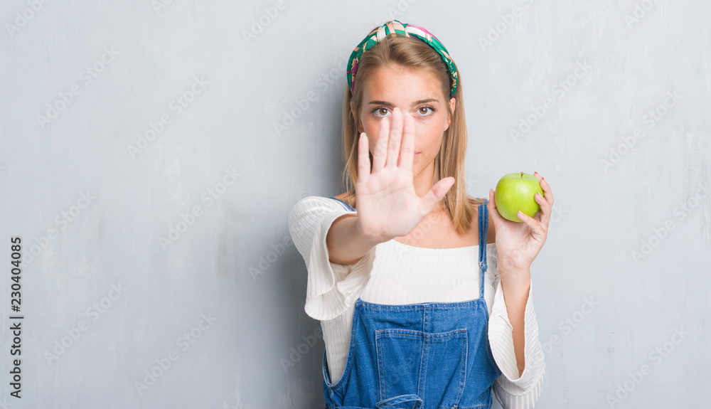 Beautiful young woman over grunge grey wall eating green apple with open hand doing stop sign with serious and confident expression, defense gesture Beautiful young woman over grunge grey wall eating green apple with open hand doing stop sign with serious and confident expression, defense gesture