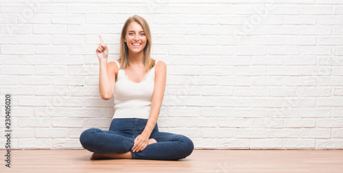 Beautiful young woman sitting on the floor at home showing and pointing up with finger number one while smiling confident and happy.