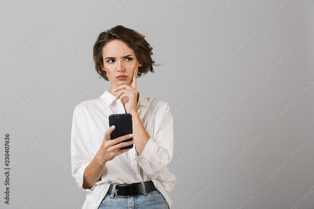Thinking confused young business woman posing isolated over grey wall background using mobile phone chatting. Thinking confused young business woman posing isolated over grey wall background using mobile phone chatting.