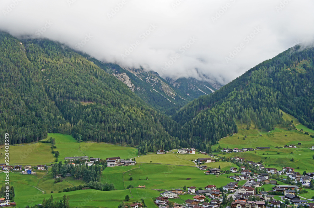 Dorf im Stubaital/ Blick auf ein Dorf im Stubaital in Tirol, Oesterreich; grünes Tal, Häuser und bewaldete Berghänge; Gipfel in Wolken.