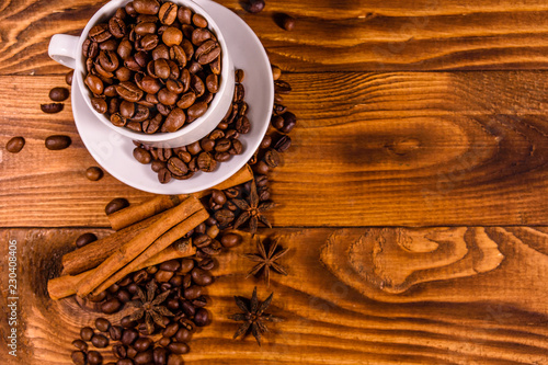 White cup filled with coffee beans, star anise and cinnamon sticks on wooden table. Top view