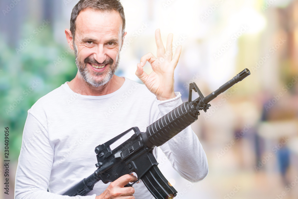 Middle age senior hoary criminal man holding gun weapon over isolated background doing ok sign with fingers, excellent symbol Middle age senior hoary criminal man holding gun weapon over isolated background doing ok sign with fingers, excellent symbol