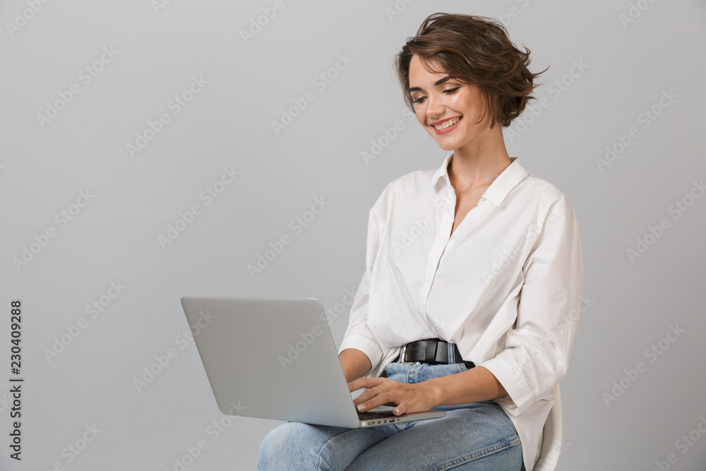 Young business woman posing isolated over grey wall background sitting on stool using laptop computer. Young business woman posing isolated over grey wall background sitting on stool using laptop computer.