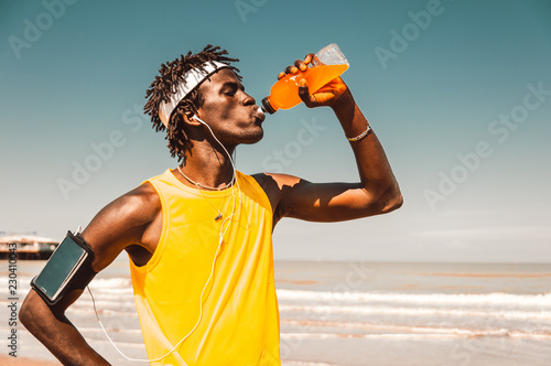 Black runner taking rest drinking energy drink at the beach at summer