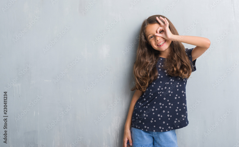 Young hispanic kid over grunge grey wall doing ok gesture with hand smiling, eye looking through fingers with happy face. Young hispanic kid over grunge grey wall doing ok gesture with hand smiling, eye looking through fingers with happy face.