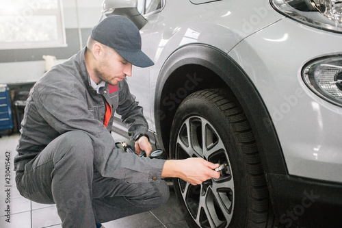 Young man in grey uniform sits in squad position in front of wheel. He looks at it and repairs tire. Guy is serious nad concentrated.