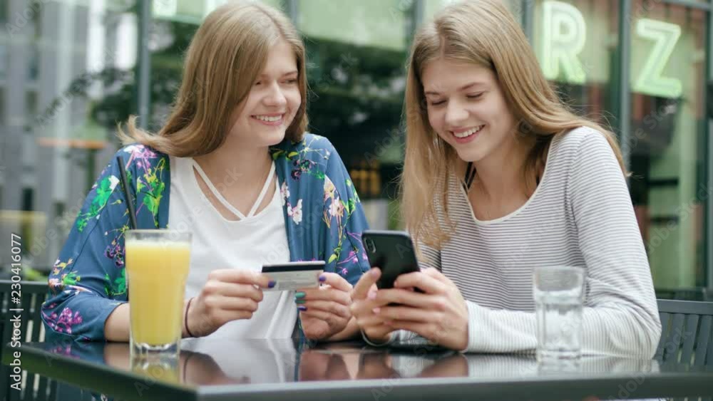 Attractive young ladies sitting at the table outdoors and using a phone and holding a credit card. Medium shot. Soft focus Attractive young ladies sitting at the table outdoors and using a phone and holding a credit card. Medium shot. Soft focus