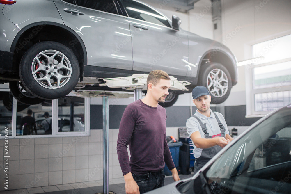 Two young men stand in garage at car. Worker points on automobile. Owner looks at it. They are serious and concentrated. There is another car on platform behind them. Two young men stand in garage at car. Worker points on automobile. Owner looks at it. They are serious and concentrated. There is another car on platform behind them.