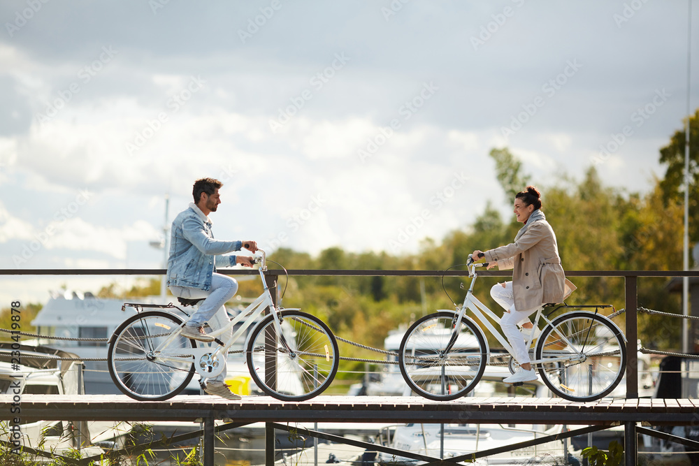 Obraz premium Cheerful young couple riding bikes directly to each other on narrow wooden pier and smiling, they meeting at seaport