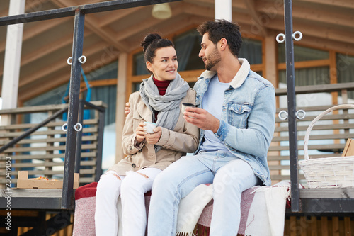 Smiling beautiful young couple in love sitting on porch of modern house and looking at each other while drinking hot tea together and enjoying picnic outdoors