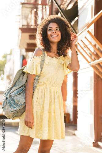 Full length photo of cheerful african american woman 20s wearing straw hat and dress, walking outdoor during sunny day