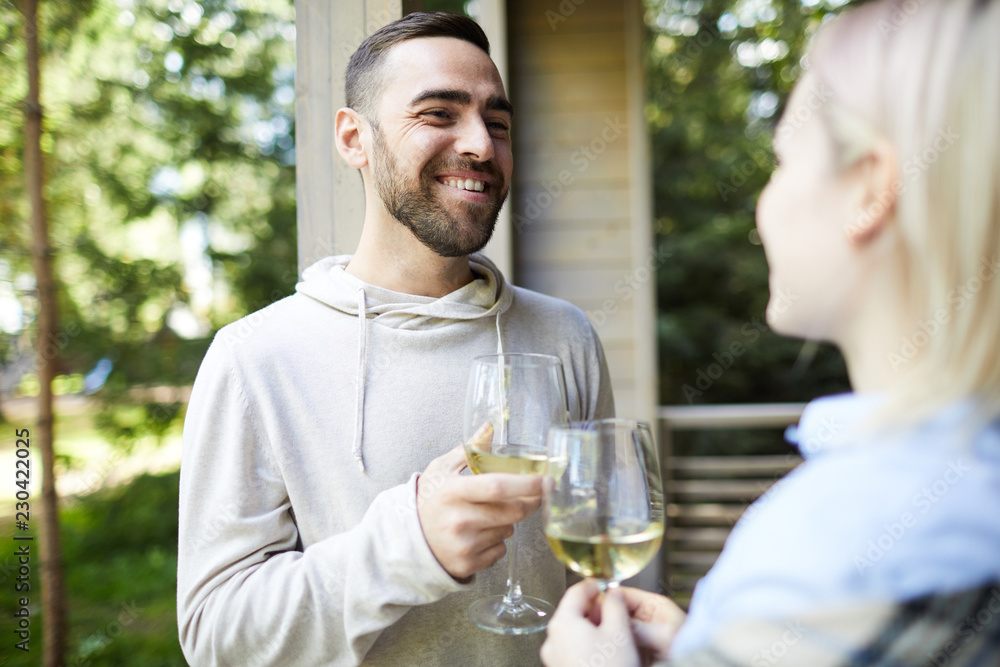 Young bearded Caucasian man smiling cheerfully while standing on house porch and clinking glasses of wine with his girlfriend Young bearded Caucasian man smiling cheerfully while standing on house porch and clinking glasses of wine with his girlfriend