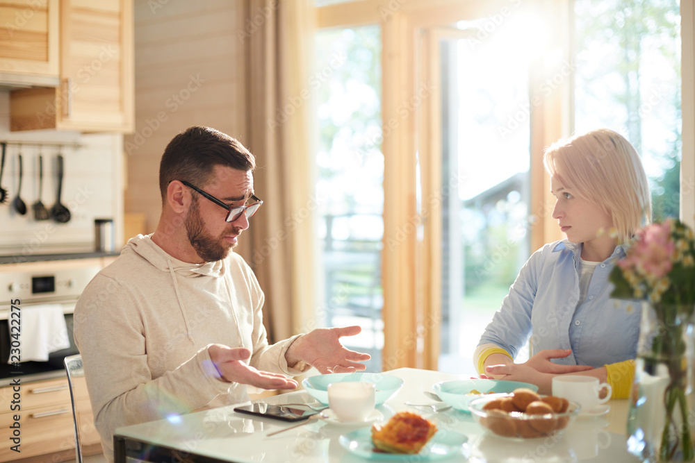 Young married Caucasian couple having serious talk while sitting at kitchen table and having breakfast on Sunday morning Young married Caucasian couple having serious talk while sitting at kitchen table and having breakfast on Sunday morning