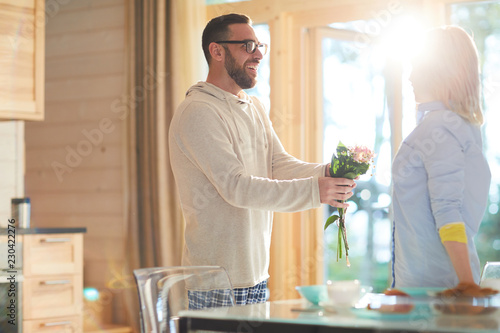 Cheerful bearded Caucasian man presenting small pretty bouquet of flowers to his girlfriend while standing in bright sunlit kitchen