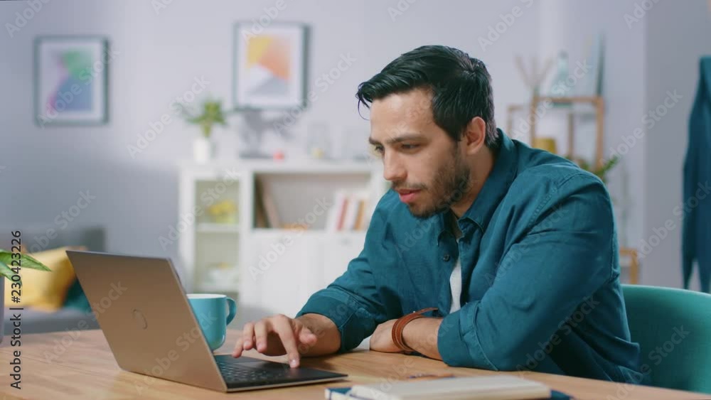 Portrait of Focused Young Man Working on a Laptop from Home, He Drinks Beverage From the Mug and is Satisfied with His Work. Cozy Modern Flat done in Scandinavian Style. Portrait of Focused Young Man Working on a Laptop from Home, He Drinks Beverage From the Mug and is Satisfied with His Work. Cozy Modern Flat done in Scandinavian Style.