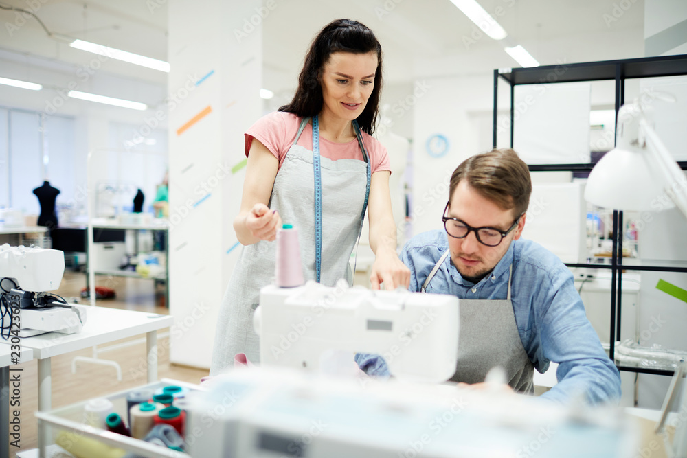 Content confident attractive young woman in apron standing at interns workplace and explaining student how to work on sewing machine in workshop Content confident attractive young woman in apron standing at interns workplace and explaining student how to work on sewing machine in workshop