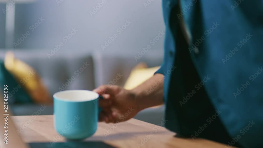Anonymous Man Enters His Living Room with a Mug and Sits Down at His Desk Starts Working on a Laptop. Anonymous Man Enters His Living Room with a Mug and Sits Down at His Desk Starts Working on a Laptop.