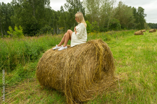 A pretty blonde woman in a In a white knitted woolen jacket sits on a large round stack of dry hay collected on a summer or autumn day against a background of green grass at half a turn position