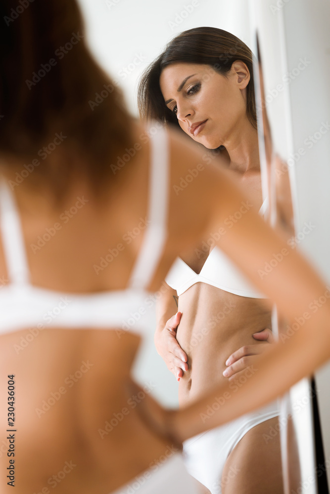 Fototapeta premium Young beautiful woman in white underwear looking herself worried about her body at the mirror in bedroom.