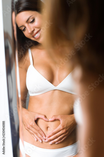 Young beautiful woman making heart shape with her hands on her belly in front of the mirror.