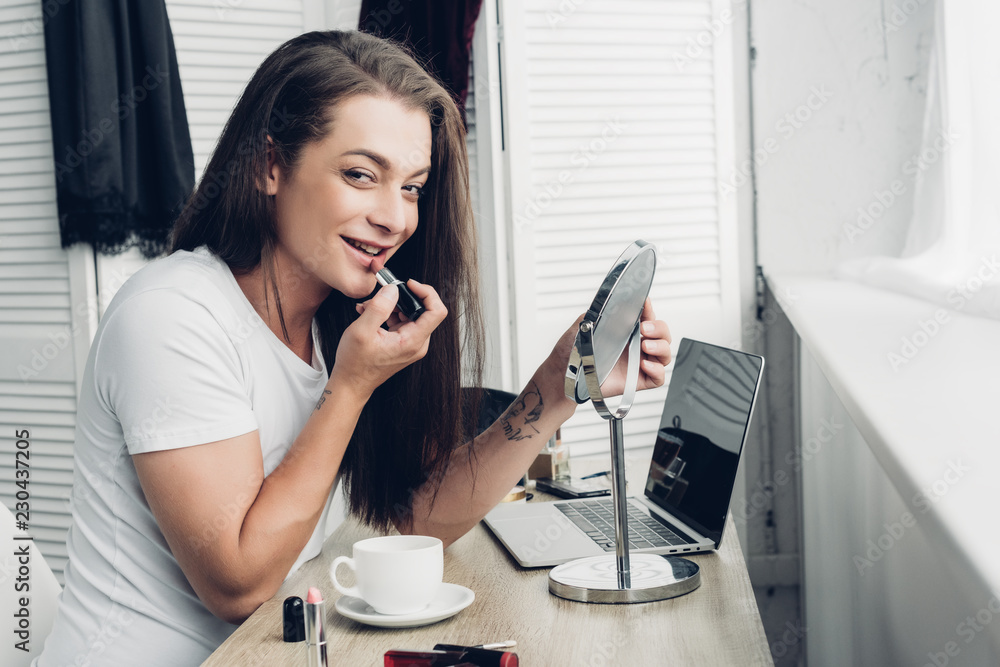 smiling young transgender man with round makeup mirror applying lipstick while looking at camera smiling young transgender man with round makeup mirror applying lipstick while looking at camera