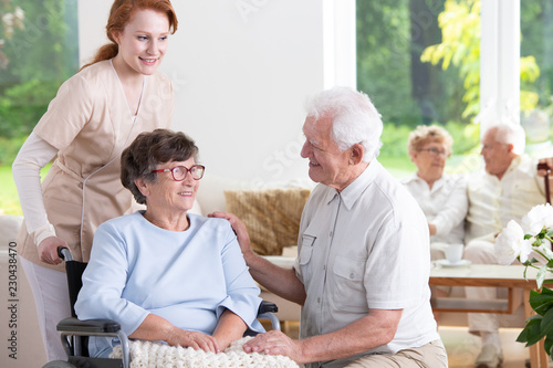 Smiling elderly man visiting senior woman in the wheelchair while nurse supporting her