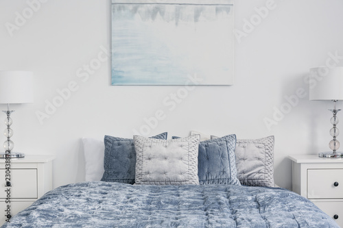 Poster above blue bed with pillows in white simple bedroom interior with lamps on cabinets. Real photo