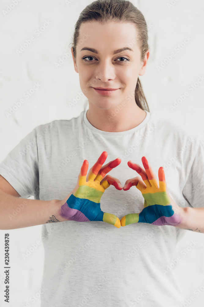 smiling young transgender man making heart sign with hands in colors of pride flag in front of white brick wall smiling young transgender man making heart sign with hands in colors of pride flag in front of white brick wall