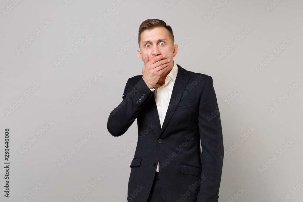 Portrait of shocked young business man in classic black suit, shirt covering mouth with hand isolated on grey wall background in studio. Achievement career wealth business concept. Mock up copy space. Portrait of shocked young business man in classic black suit, shirt covering mouth with hand isolated on grey wall background in studio. Achievement career wealth business concept. Mock up copy space.