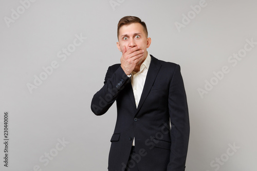 Portrait of shocked young business man in classic black suit, shirt covering mouth with hand isolated on grey wall background in studio. Achievement career wealth business concept. Mock up copy space.