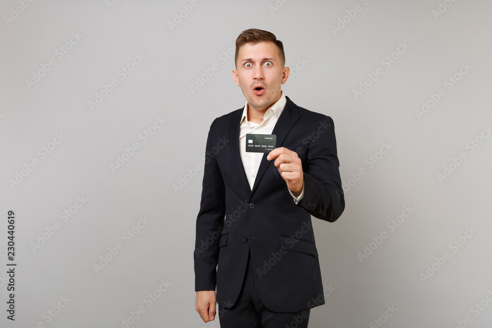 Shocked young business man in classic black suit and shirt keeping mouth wide open holding credit bank card isolated on grey background. Achievement career wealth business concept. Mock up copy space. Shocked young business man in classic black suit and shirt keeping mouth wide open holding credit bank card isolated on grey background. Achievement career wealth business concept. Mock up copy space.