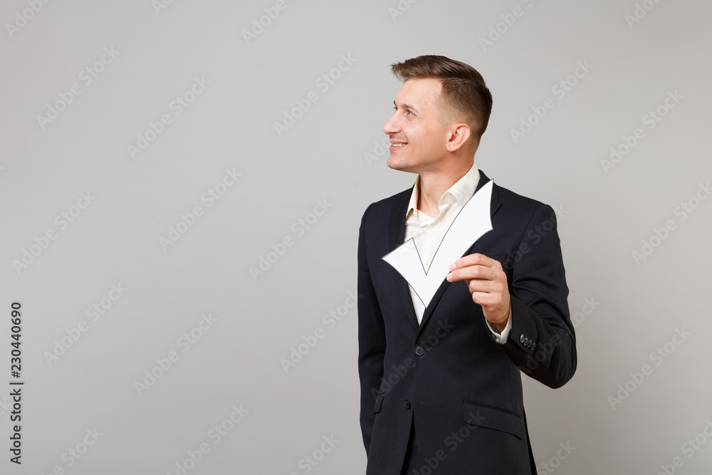 Portrait of young business man in classic black suit, shirt looking aside holding check mark isolated on grey wall background in studio. Achievement career wealth business concept. Mock up copy space. Portrait of young business man in classic black suit, shirt looking aside holding check mark isolated on grey wall background in studio. Achievement career wealth business concept. Mock up copy space.