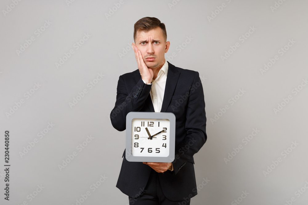 Upset young business man in classic black suit, shirt putting hand on cheek, holding square clock isolated on grey background in studio. Achievement career wealth business concept. Mock up copy space. Upset young business man in classic black suit, shirt putting hand on cheek, holding square clock isolated on grey background in studio. Achievement career wealth business concept. Mock up copy space.