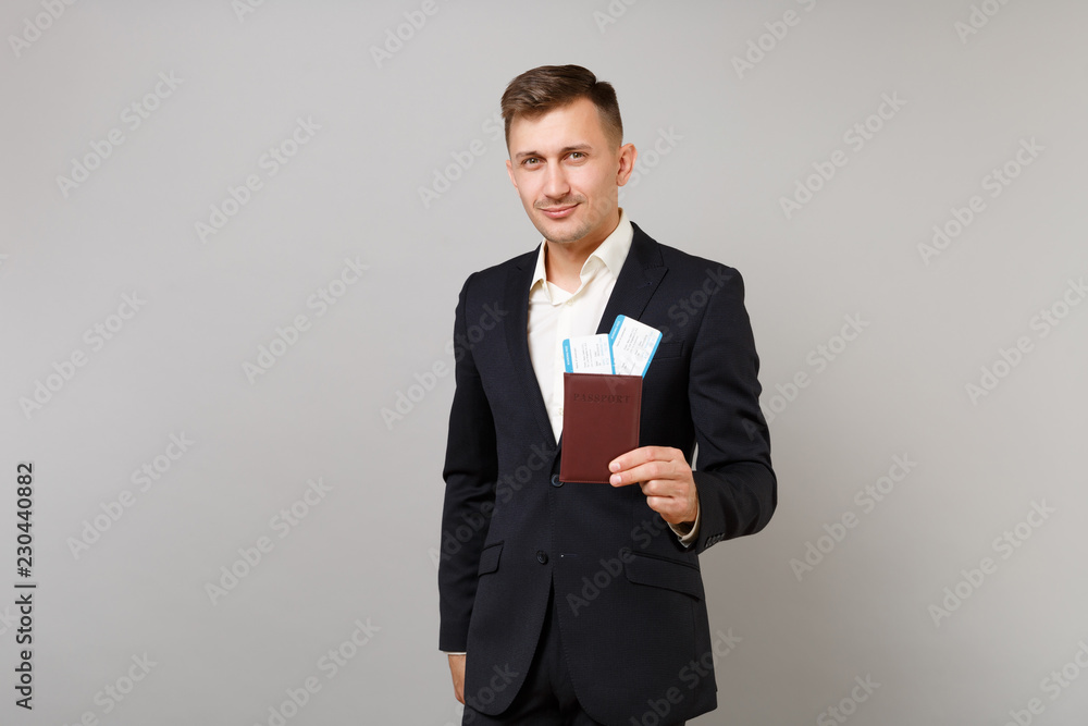 Handsome young business man in classic black suit, shirt hold passport, boarding pass ticket isolated on grey wall background in studio. Achievement career wealth business concept. Mock up copy space. Handsome young business man in classic black suit, shirt hold passport, boarding pass ticket isolated on grey wall background in studio. Achievement career wealth business concept. Mock up copy space.
