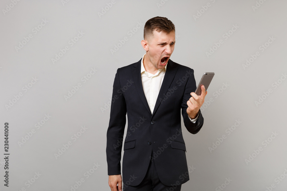 Irritated business man in classic black suit screaming, looking on mobile phone in hand, making video call isolated on grey background. Achievement career wealth business concept. Mock up copy space. Irritated business man in classic black suit screaming, looking on mobile phone in hand, making video call isolated on grey background. Achievement career wealth business concept. Mock up copy space.