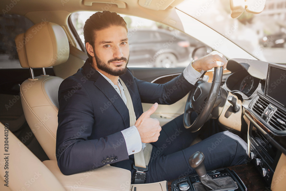 Nice picture of young businessman sitting in luxury car and holding big thumb up. He hold one hand on steering wheel. Young man recommends this car. Nice picture of young businessman sitting in luxury car and holding big thumb up. He hold one hand on steering wheel. Young man recommends this car.